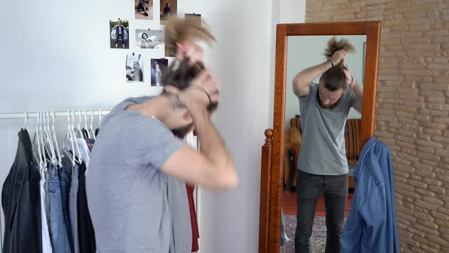 Attractive Young Caucasian beard man in a T-shirt  is looking in a mirror tying his hair 