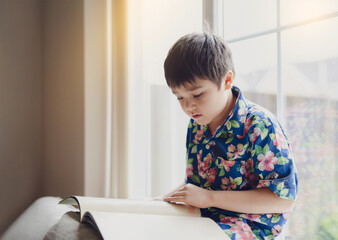 Naklejka premium Authentic Kid reading a book with bright light shining in the morning,Happy boy sitting next to window reading a story or cartoon, Hight key light Child siting on sofa relaxing at home on Summer