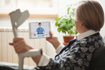 Retired senior elderly woman holding crutch and tablet computer talking to NHS GP orthopedic female...