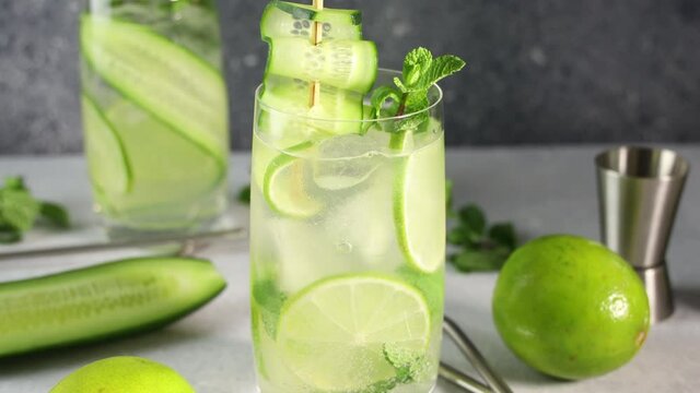 Decoring mojito cocktail or detox cocktail of mint, cucumber and lime in highball glass on a gray concrete stone surface background. Camera moving along glass.