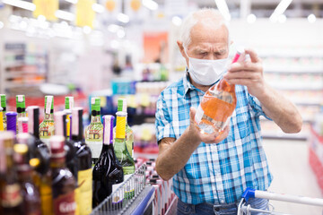 mature european man wearing mask with covid protection chooses bottle of alcohol in supermarket