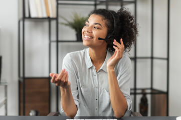 Young smiley African American female customer service agent in a headset, touching microphone,...