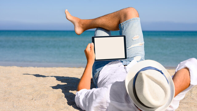 A man in a white shirt and hat is lying on the beach with a tablet in his hands. Blank white display on tablet.