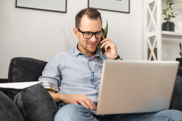 A smiling freelancer guy wearing glasses solving tasks by smartphone working with a laptop from home