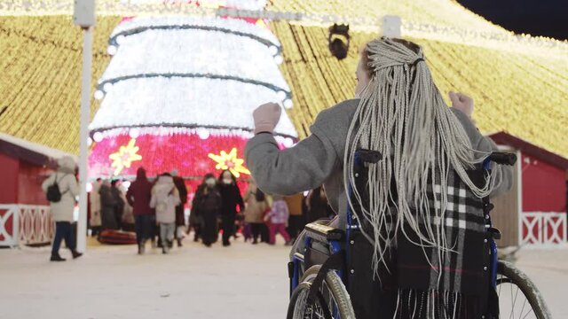A young woman with grey dreadlocks in a wheelchair looks at a Christmas tree and crowds of people and slightly dancing