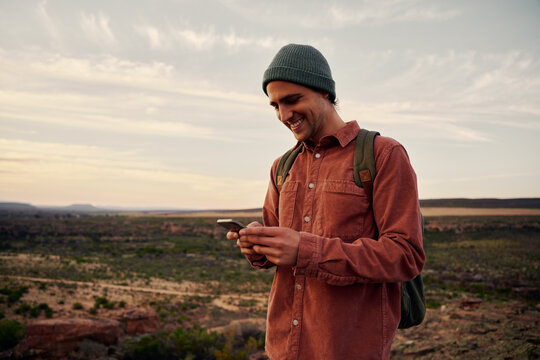 Happy male hiker with backpack and cap using smartphone on mountain