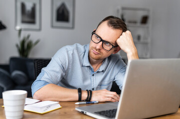 An young man is bored working remotely from home, a guy is holding head and looks away