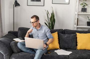 Thoughtful and intelligent entrepreneur looking through notes sitting on the comfortable couch with laptop. A freelancer wearing glasses works online remotely