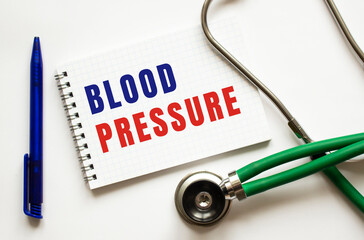 BLOOD PRESSURE is written in a notebook on a white table next to pen and a stethoscope.