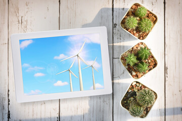 Wind turbine clean energy on fluffy cloud and blue sky background on computer tablet with cactus on desk background. Clean energy to sustainable future concept and alternative energy idea