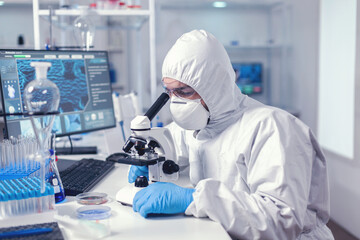 Healthcare specialist analyzing vaccine on microscope dressed in protection suit. Virolog in...