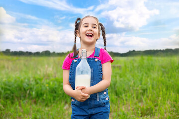 A joyful laughing girl holds a glass bottle of milk in summer, in a field on the grass.