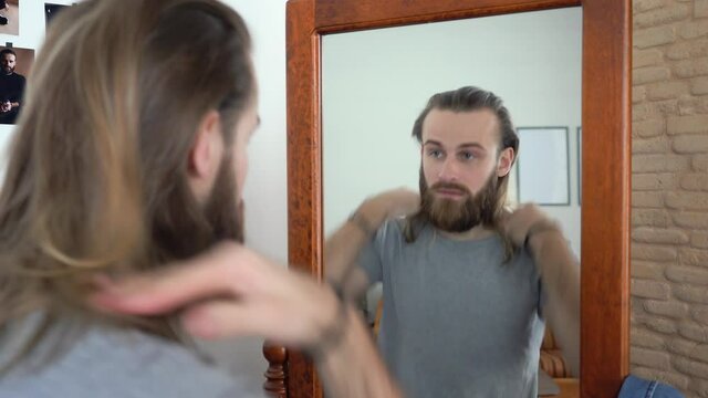 Attractive Young Brutal beard man in a T-shirt  is looking in a mirror twist out his hair 