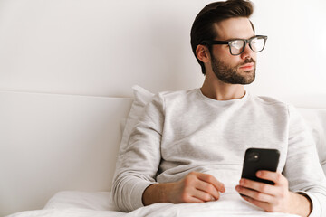 Serious young man in eyeglasses using mobile phone while resting in bed