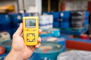 Using a portable gas detector to monitor the air or toxic gas composition around the chemical storage area (with blurred background of chemical barrel). Industrial action and object photo. - obrazy, fototapety, plakaty