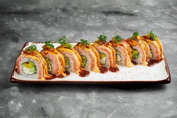 Japanese sushi roll with smoked salmon, scrambled eggs, avocado and cream cheese topped with unagi and spicy sauce. Sushi in white plate on gray background. Copy space