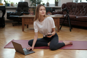 Fototapeta na wymiar Portrait of a fitness girl with telephone using laptop which sits on mat around luxurious furniture and modern photocamera.