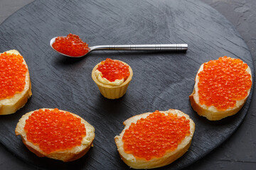 Canapes with butter and red caviar and a spoon with caviar on a black round board on a concrete background copy space