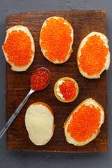 Bruschetta with butter and red caviar next to a spoon with caviar on a board on a concrete background.