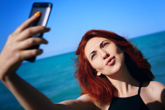 Red-haired girl makes her lips duck and takes selfie on smartphone camera. Summer sunny vacation. Pretty woman in black swimsuit takes pictures of herself against background of sea and sky.