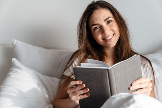 Happy brunette nice girl reading book while sitting in bed at home