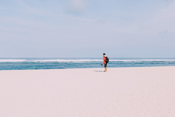 Lonely man in red T-shirt walking on white beach against blue ocean. Traveling alone.