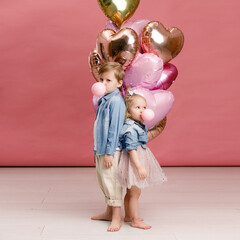 Little boy and girl hugging, dancing, smiling and having fun near big branch of pink heart-shaped ballons. Valentin’s day concept . Isolated on pink background.