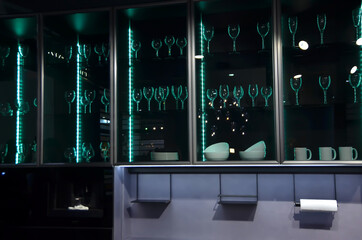 Modern kitchen in black minimalist style at night with LED green ribbon. There are glasses on the shelves behind the glass