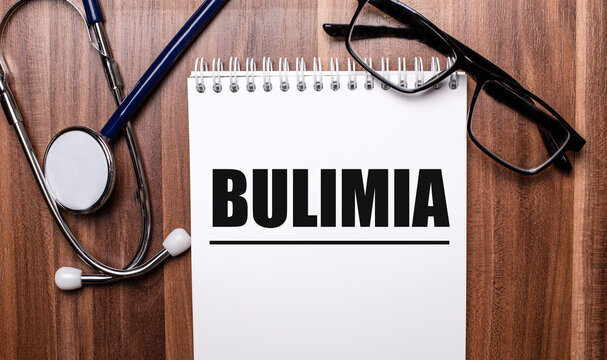The word BULIMIA is written on white paper on a wooden background near a stethoscope and black-framed glasses. Medical concept
