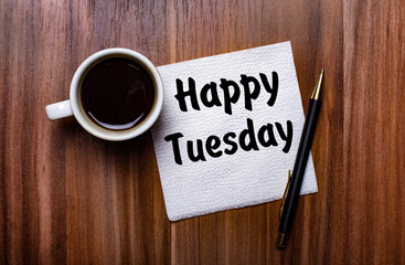 On a wooden table next to a white cup of coffee and a pen is a white paper napkin with the words HAPPY TUESDAY