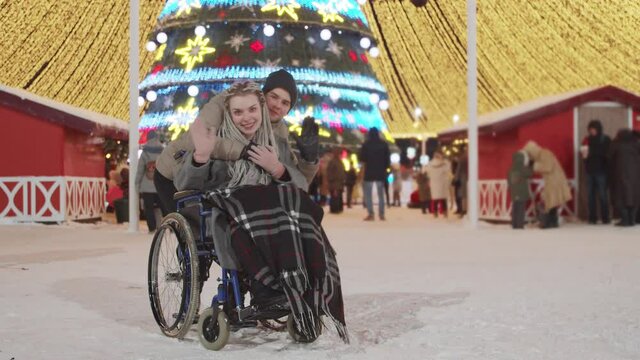Smiling young woman in a wheelchair and her boyfriend at Christmas party on the streets - waving hands to the camera