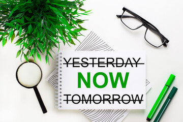 On a white background lies a notebook with the word YESTERDAY NOW TOMORROW, glasses, a magnifying glass, green markers and a green plant
