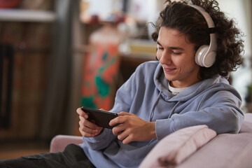 Teenage boy in wireless headpones talking online using his smart phone sitting on sofa