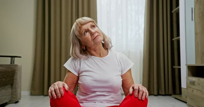 Old beautiful woman grandmother independently performs exercises for neck rehabilitation after injury at home sitting on the ground in sportswear