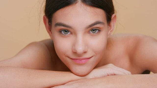 Close-up beauty portrait of young attractive brunette woman raises her head from her forearm, laying chin on her hands, looks at camera and smiles