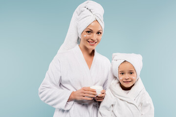 happy mother in bathrobe holding container with face cream near daughter isolated on blue