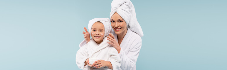 happy mother in bathrobe applying cosmetic cream on face of daughter isolated on blue, banner
