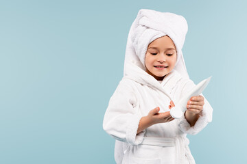 smiling child in bathrobe and towel on head holding tube with cosmetic product isolated on blue