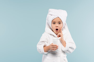 shocked kid in bathrobe and towel on head holding tube with cosmetic product isolated on blue