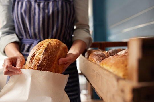 Sales Assistant In Bakery Putting Freshly Baked Organic Sourdough Bread Loaf Into Sustainable Paper Bag