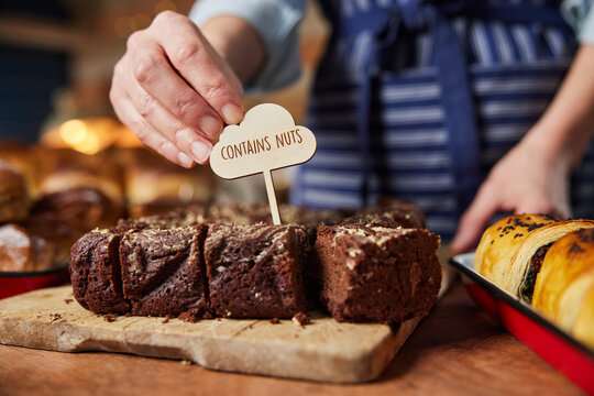 Sales Assistant In Bakery Putting Contains Nuts Label Into Stack Of Freshly Baked Baked Brownies