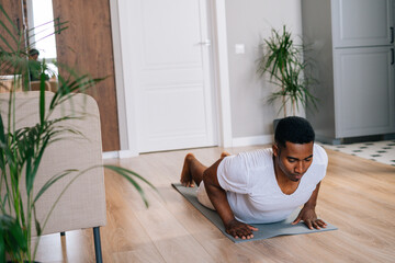 Strong African-American man practicing burpee exercise at home, doing push-ups and jumping on yoga mat at bright domestic room. Concept of sport training at home gym.