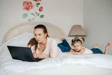 Work from home with kids children. Mother working on laptop in bedroom with child daughter toddler beside her. Funny candid family moment. New normal during coronavirus quarantine, lockdown.