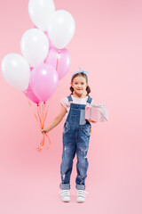 full length of cheerful kid holding balloons and gift box on pink