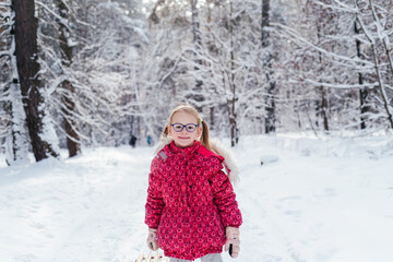 Little girl in eyeglasses pulls sled through beautiful winter forest
