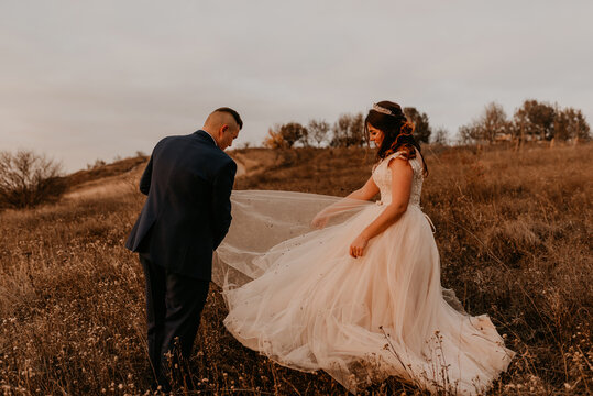 Groom Cleans Mud From Bride Wedding Dress In Middle Of Field In Tall Grass In Autumn