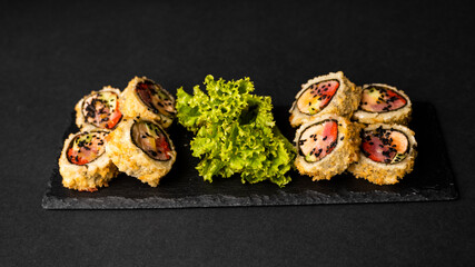 Custom sushi roll in tempura with nori, fresh salmon, tuna, avocado, masago caviar, drizzled with pineapple sauce with salad pouring as decoration on a black plate on a black table and background.