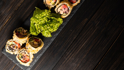 Custom sushi roll in tempura with nori, fresh salmon, tuna, avocado, masago caviar, drizzled with pineapple sauce with salad pouring as decoration on a black plate on a wooden table and background.