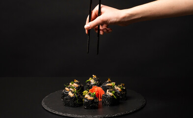 Hand with chopsticks wants to take custom sushi roll with black rice, crab meat, avocado, smoked salmon mousse, oar caviar, masago, shrimp cocktail, edible gold leaf, ginger, wasabi on black table.