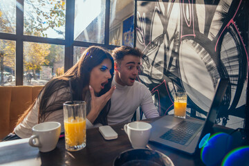 Cute couple are looking surprised at their laptop while sitting in a urban cafe near a window